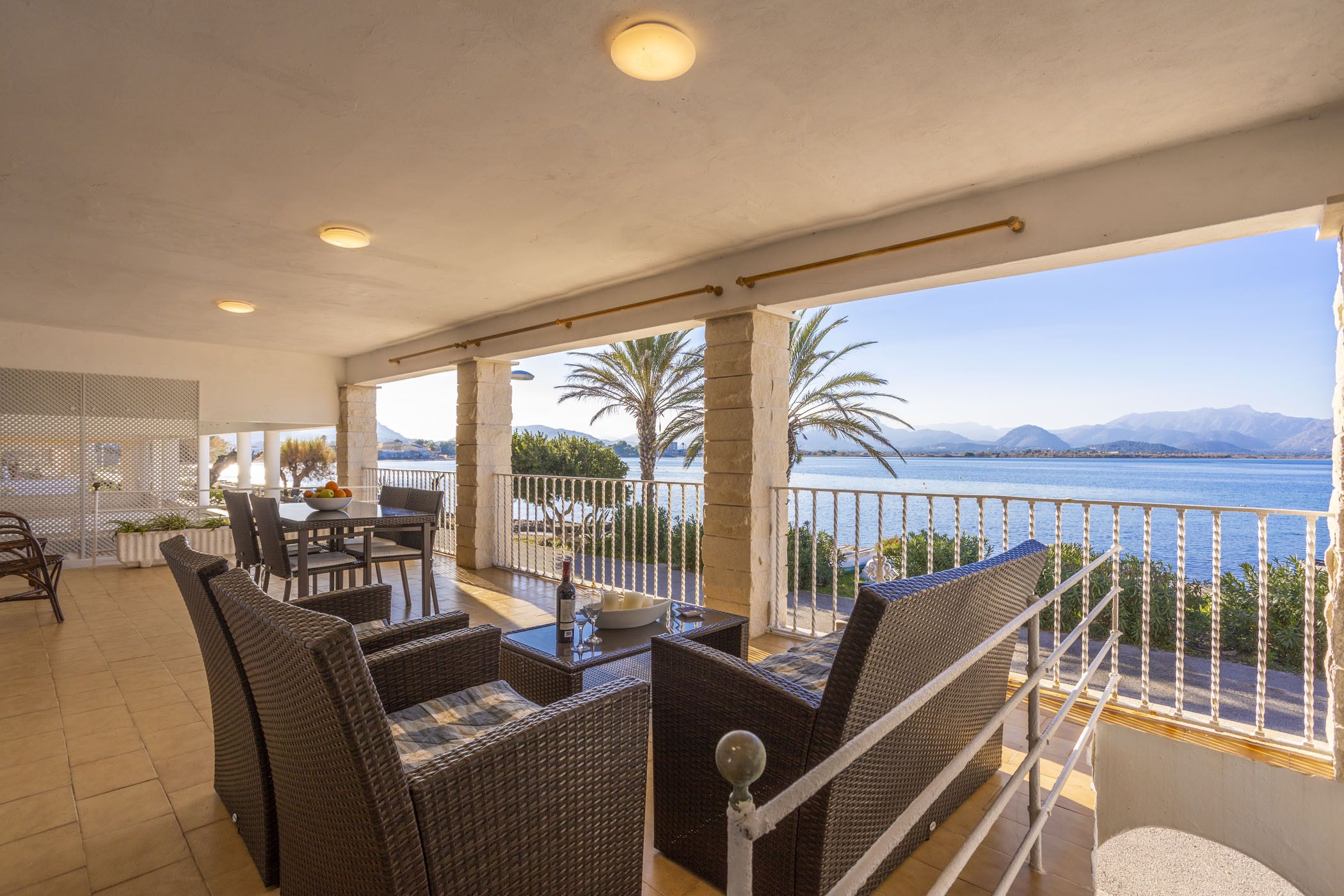 Un balcon agrémenté de meubles en osier, une table avec une bouteille, et une vue sur la mer et les montagnes.
