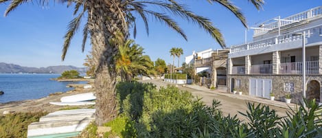 A coastal building with a balcony overlooking the sea, surrounded by greenery and palm trees.