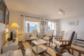 A hotel room with a view of the sea, a white coffee table, two white armchairs, a wooden chair, a small white table with a candle, and a lamp.