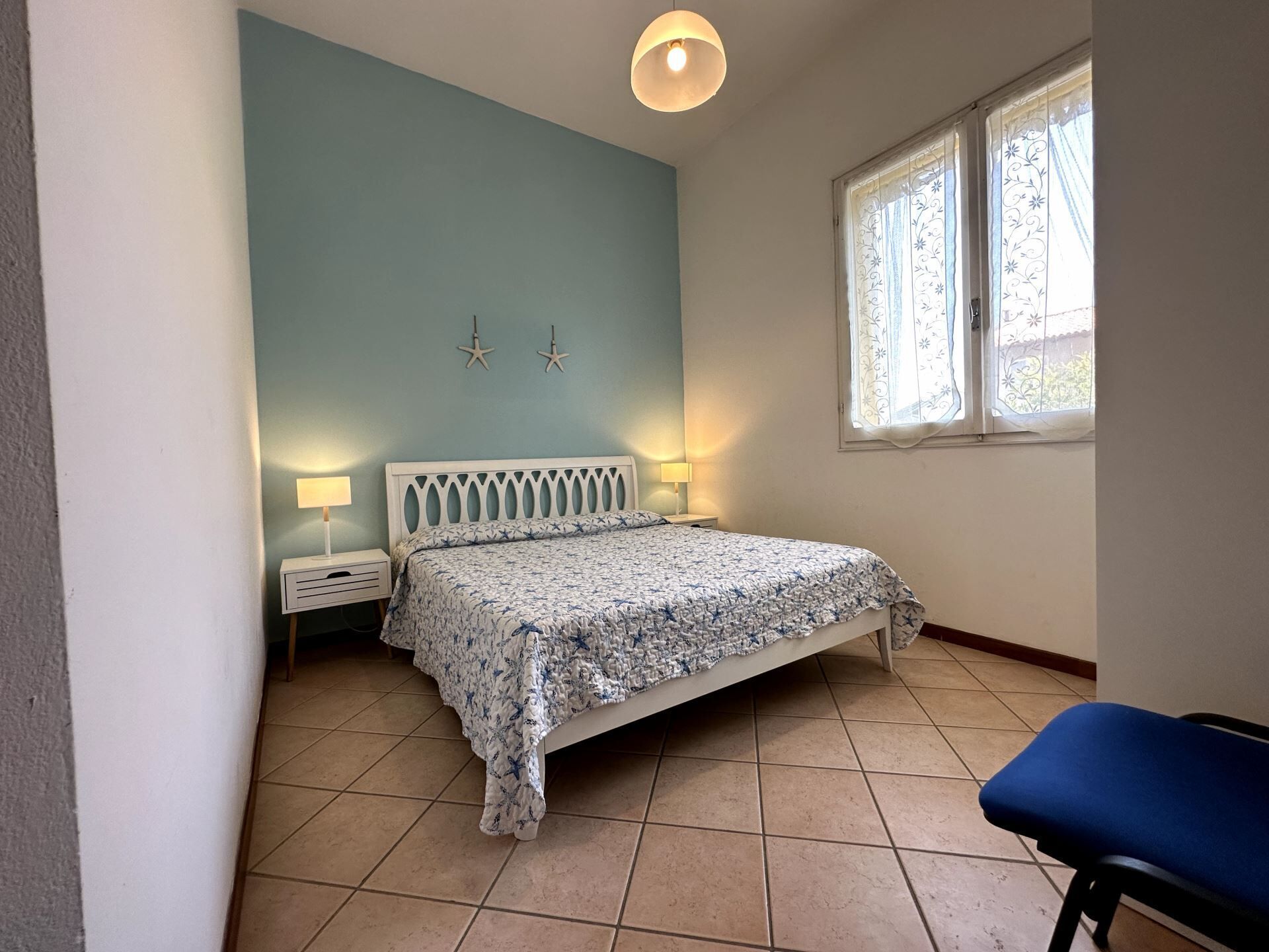 A bedroom with a bed, two bedside lamps, a window with curtains, and a blue chair.