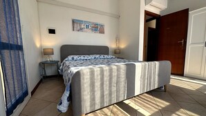 A bedroom with a bed, a nightstand, a wall-mounted lamp, a door, and a window with curtains. - Delta Blu Residence Village - Appartamento Quadrilocale 2 (Lido di Pomposa)