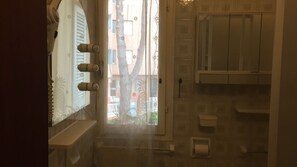 Ein Badezimmer mit Dusche, Toilette und Waschbecken. Ein Fenster mit Jalousien und eine Tür zu einem Außenbereich.