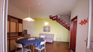 A dining area with a round table, chairs, a hanging light fixture, a staircase, and a framed picture on the wall. - Delta Blu Residence Village - Villetta Trilocale 19 (Lido di Pomposa)