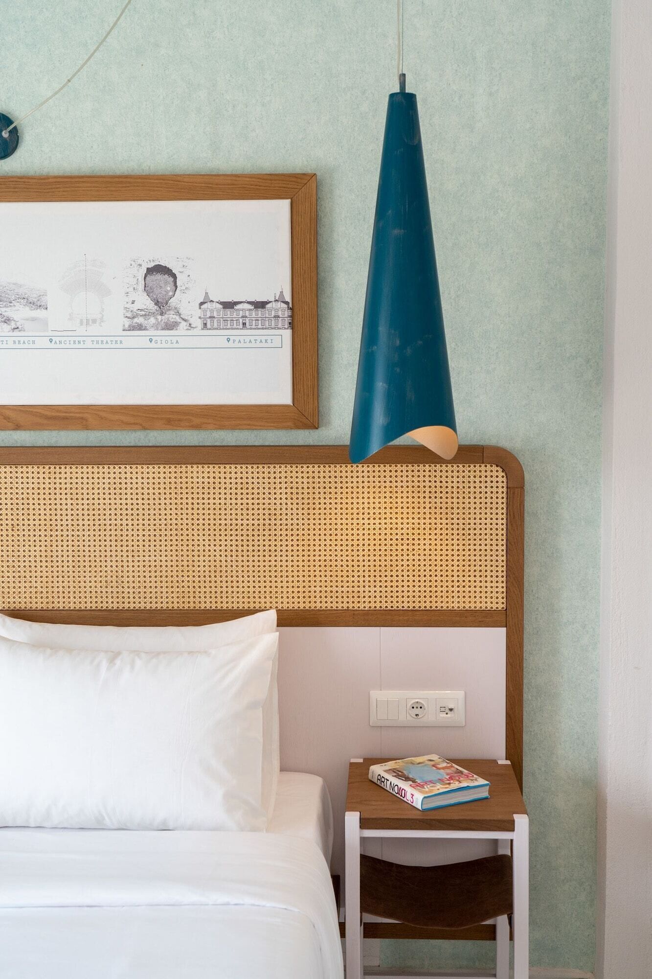 Une chambre à coucher avec un lit, une table de chevet sur laquelle repose un livre, une carte encadrée et une lampe suspendue bleue.