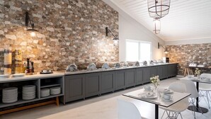 A modern kitchen with a stone wall backsplash, a long counter with various kitchenware, and a dining area with tables and chairs. - Tripiti Bungalows (Thasos)