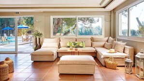 A spacious living area with a beige sectional sofa, a matching ottoman, and a small round table. Large windows provide a view of the outdoor pool area. - Tripiti Bungalows (Thasos)