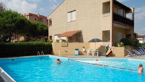 A swimming pool with clear blue water, surrounded by a building with a balcony and a small patio area with a table and chairs. - Delta Blu Residence Village - Villetta Trilocale 10 (Lido di Pomposa)