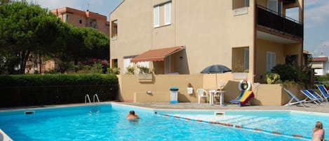 Una piscina con agua cristalina azul, rodeada por un edificio con balcón y un pequeño patio con mesa y sillas.