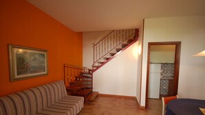 A room with a wooden staircase, a striped sofa, and a kitchen area visible through an open door. - Delta Blu Residence Village - Villetta Trilocale 10 (Lido di Pomposa)