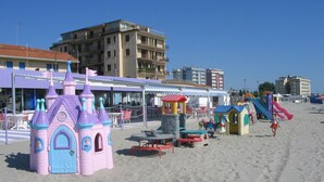 Een strand met kleurrijke speeltoestellen, waaronder een kasteel en glijbanen, en een gebouw met terrasjes op de achtergrond.