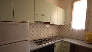 Een keuken met een witte koelkast, een gasfornuis en een gootsteen. Boven het aanrecht zijn kastjes en er is een raam met een gordijn.
