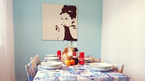 Mesa de jantar posta com pratos, uma garrafa e duas laranjas, parede azul e um quadro pendurado acima.
