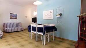 A cozy living room with a striped sofa, a small dining table with chairs, a TV, and a wall-mounted shelf with flowers.