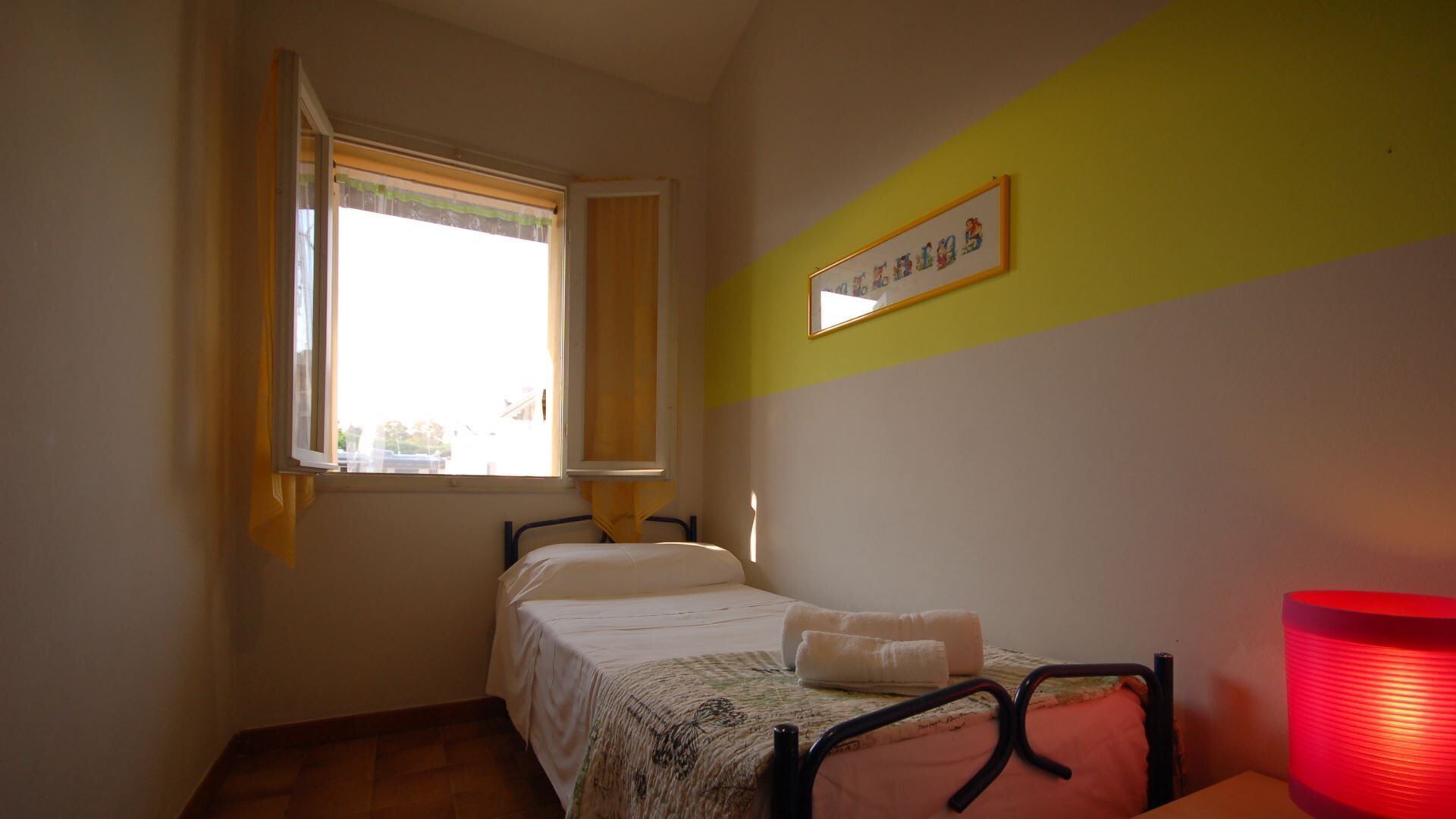 A small, single-bed room with a window, a bedside table, and a red lamp.