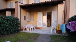 Une terrasse couverte avec une table et des chaises, un petit escalier menant à une porte, et une haie sur la gauche.