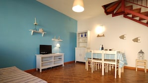 Une pièce avec un mur bleu, un escalier en bois, une table à manger avec des chaises, un téléviseur posé sur un meuble blanc et une étagère ornée d’objets décoratifs.