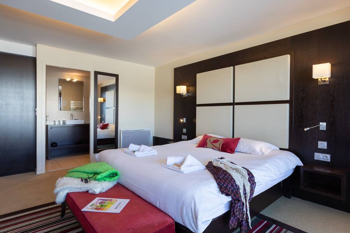 A hotel room with a large bed, a bedside table, a red bench, a mirror, a bathroom with a sink and mirror, and a wall-mounted light fixture.