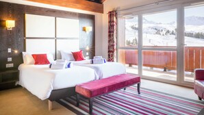Habitación de hotel con dos camas, un ventanal con vistas a la montaña, vigas de madera en el techo y un balcón con barandilla roja.