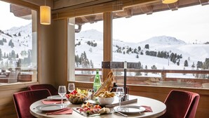 Una mesa de comedor dispuesta con platos, vasos y diversos alimentos, con vistas a montañas nevadas a través de amplios ventanales.