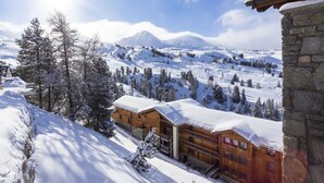 Una cabaña de madera con estructura de piedra, rodeada de árboles cubiertos de nieve y montañas al fondo.