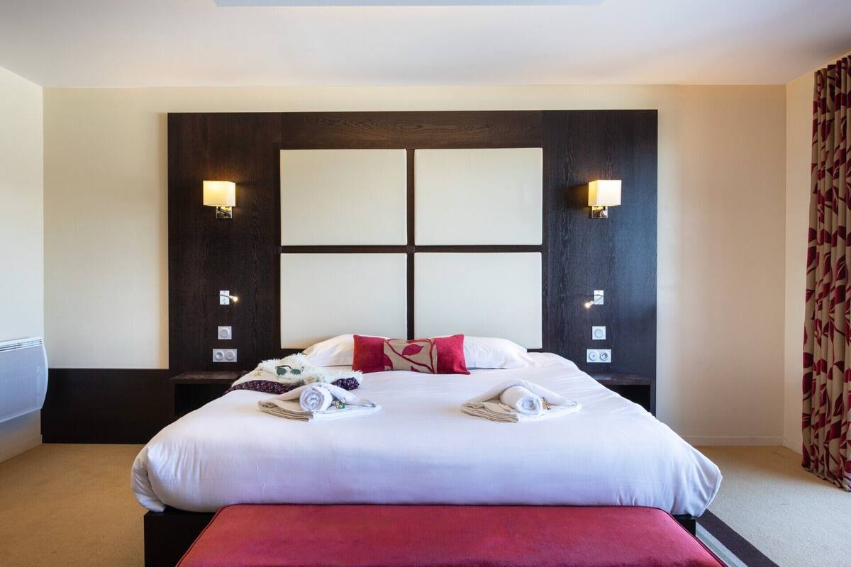 A hotel room with a large bed, two bedside lamps, a headboard with a geometric design, and a red bench at the foot of the bed.