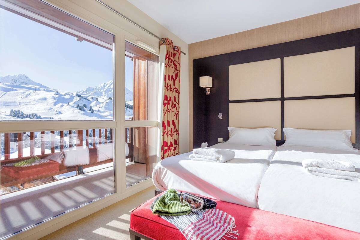 A hotel room with a large bed, a view of a snowy mountain landscape, a balcony with a wooden railing, and a red bench.
