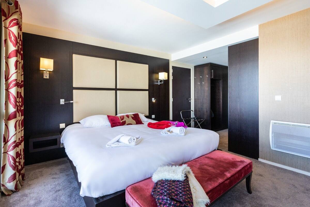 A hotel room with a large bed, a red bench, a wall-mounted heater, and a door leading to a bathroom.