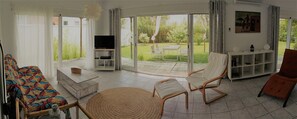 A spacious living room with a sofa, chairs, a TV, and a bookshelf. A sliding glass door leads to a garden. - Casa Montecote Playa (Caños de meca)
