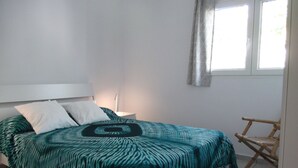 Une chambre à coucher avec un lit, des oreillers, une table de chevet, une lampe, une fenêtre avec des rideaux et une chaise.