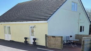 A single-story building with a sloped roof, a front door, and a small patio area with a table and chairs. - Country Retreat Sleeps 2 Near Cymyran Beach (Holyhead)
