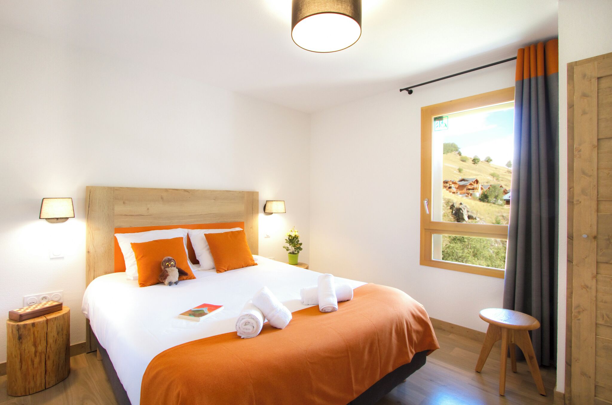 A hotel room with a wooden headboard, a large window with a view of the countryside, a bedside table with a lamp, and a small stool.