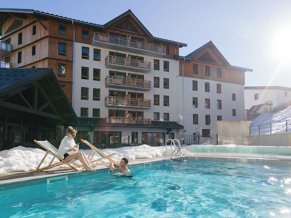 A swimming pool with a wooden deck and lounge chairs, surrounded by snow and a multi-story building with balconies.