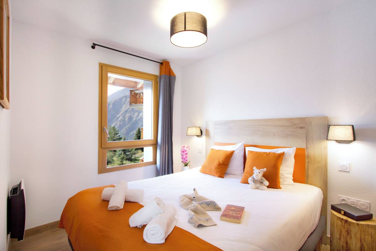 A hotel room with a large bed, a wooden headboard, a window with a view of mountains, and a nightstand with a lamp.