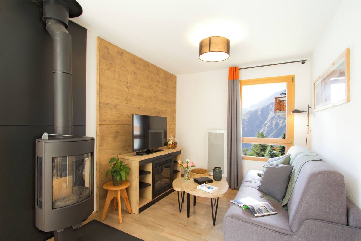 A modern living room with a fireplace, a television, a sofa, and a window with a mountain view.