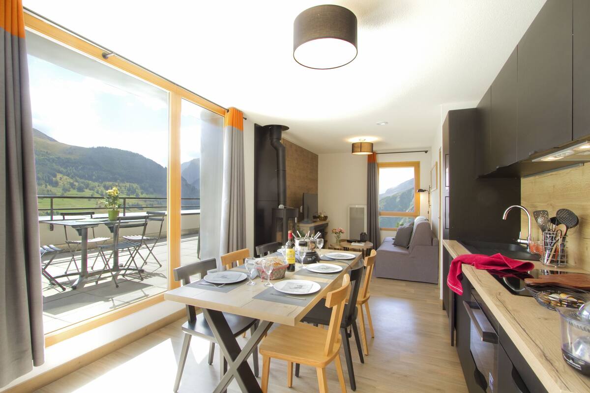 A modern dining area with a large table set for four, a kitchen with dark cabinetry, and a scenic mountain view through large windows.