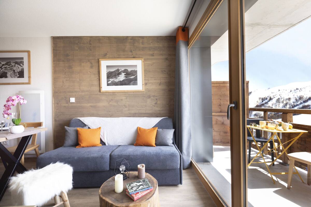 A modern living room with a grey sofa, wooden accents, a dining area with a table and chairs, and a view of a snowy landscape.