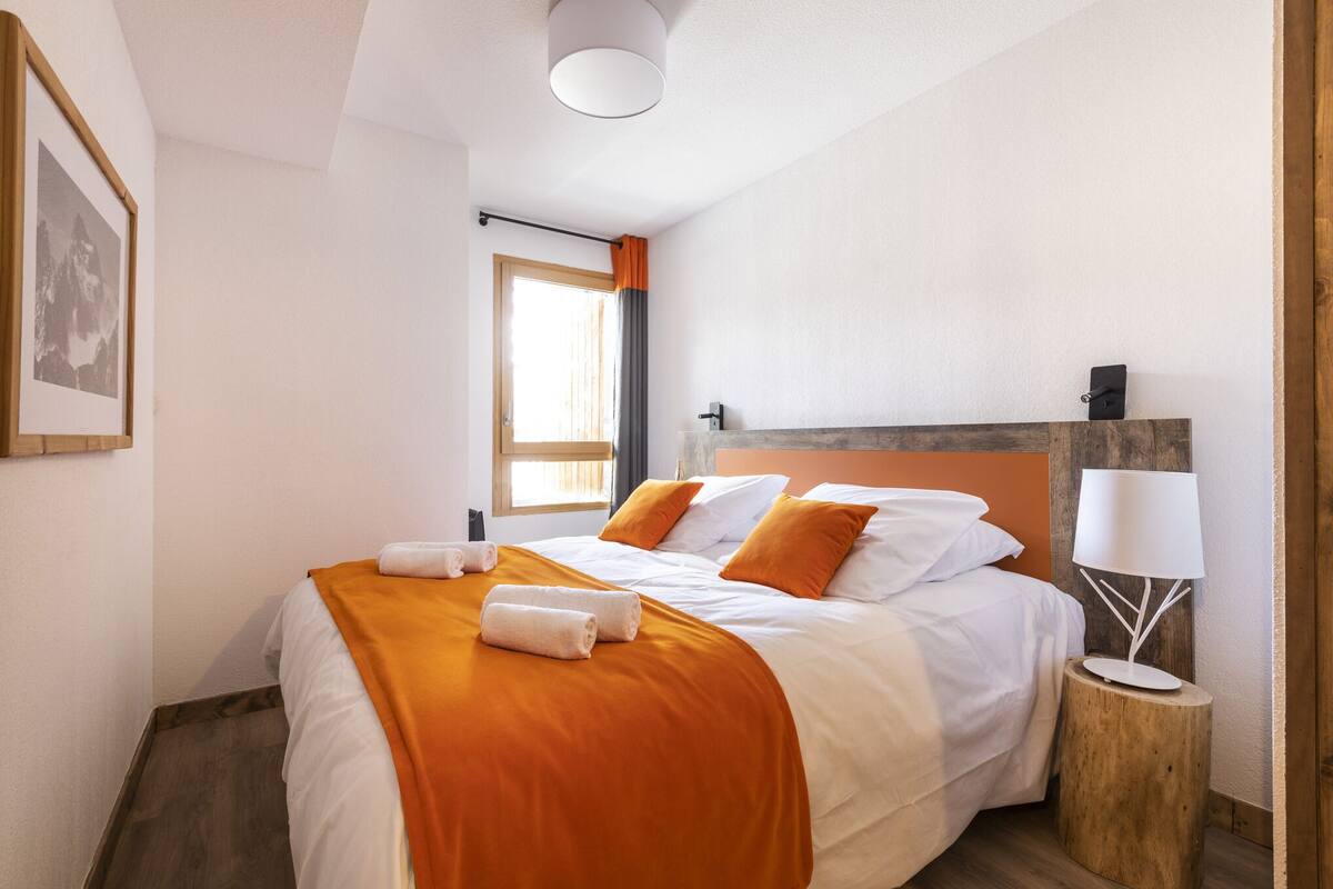 A neatly made bed with a white and orange bedspread, a wooden nightstand with a lamp, and a framed picture on the wall.