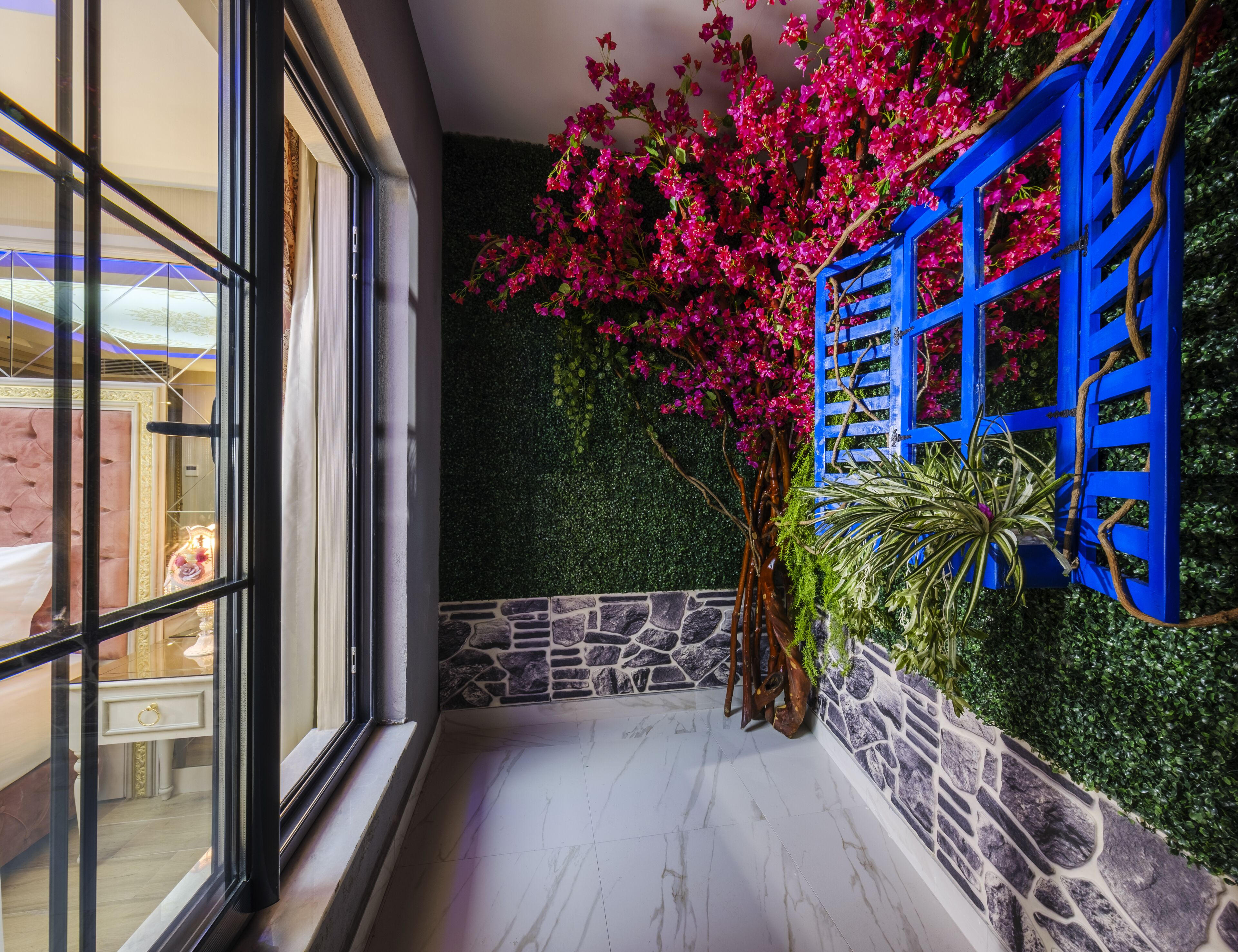 A corridor with a stone wall, a blue wooden gate, and a potted plant with pink flowers.
