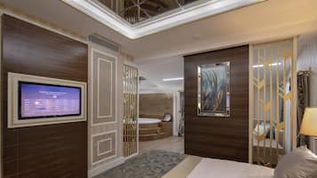 Ein modernes Hotelzimmer mit einem großen Flachbildfernseher, einem Bett mit einem gemusterten Überwurf, einer dekorativen Wandverkleidung und Blick auf einen Wellnessbereich.