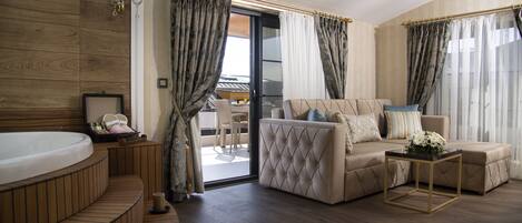 Una sala de estar moderna con un sofá, un reposapiés y una mesita con un jarrón. Hay un ventanal con cortinas y vistas al exterior.