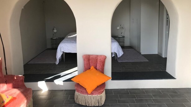 A room with two beds under arched ceilings, a small seating area with a chair and ottoman, and a visible bathroom with a mirror and towel rack.
