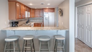 A modern kitchen with wooden cabinets, a granite countertop, a built-in stove, a refrigerator, and three bar stools. - Here Comes The Sun #4 | Oceanfront + Pet-Friendly Townhome! (Carolina Beach)