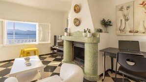 Une pièce avec un sol à carreaux, une cheminée, un ordinateur portable posé sur un bureau, une table à manger avec un saladier, et une fenêtre donnant sur un beau paysage.