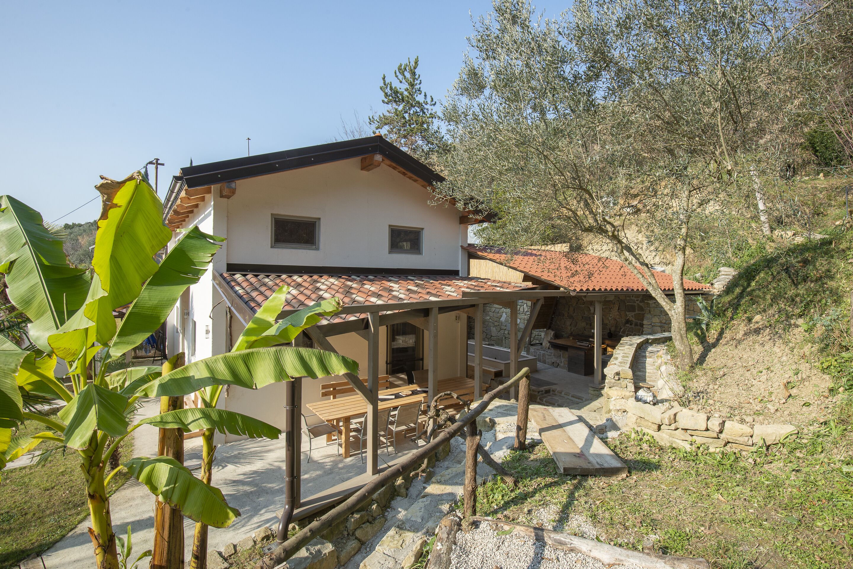 A house with a tiled roof, a covered patio with wooden furniture, and a stone pathway leading to a stone building.