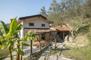 A house with a tiled roof, a covered patio with wooden furniture, and a stone pathway leading to a stone building. - Holiday Home Casa Salina (Sečovlje)