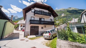 Ein zweigeschossiges Haus mit Balkon, ein geparktes Auto davor und eine Bergkulisse.
