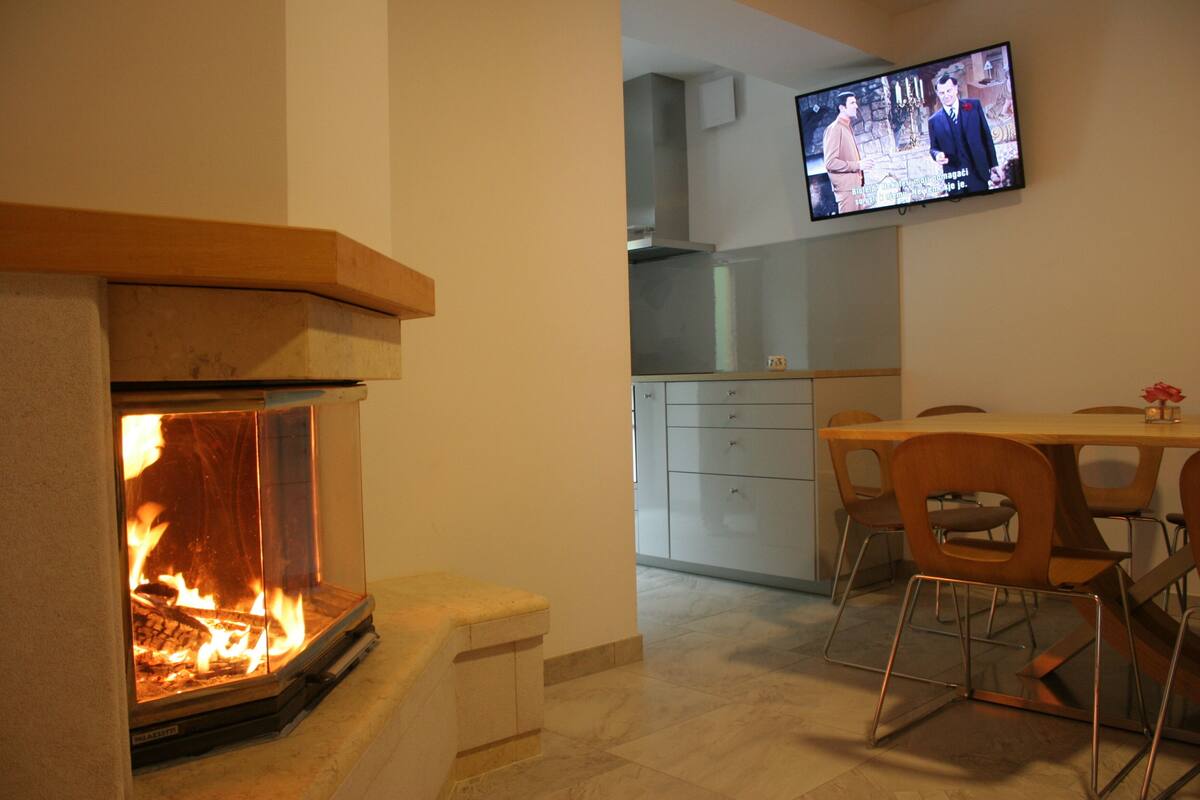 A modern living room with a fireplace, a television mounted on the wall, and a dining area with chairs and a table.
