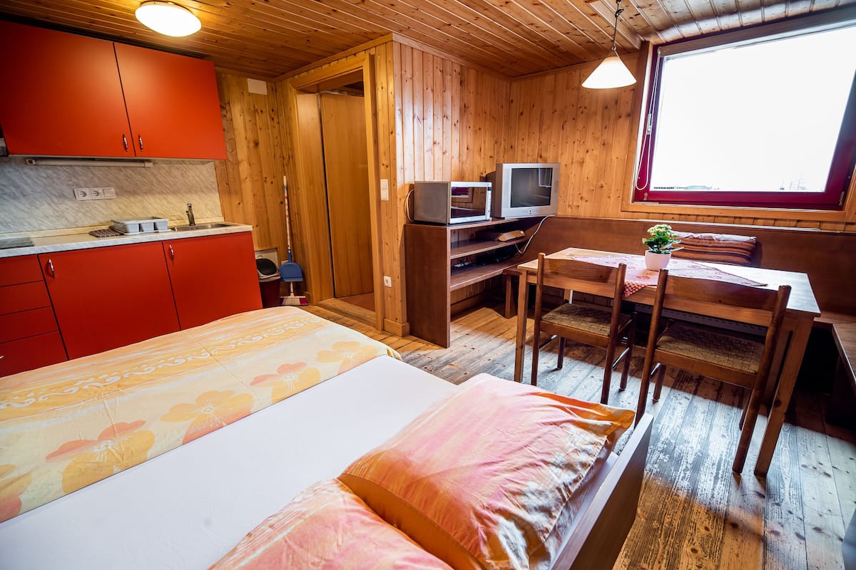 A compact room with a bed, a wooden dining table with chairs, a small kitchenette with red cabinets, and a window with a view.