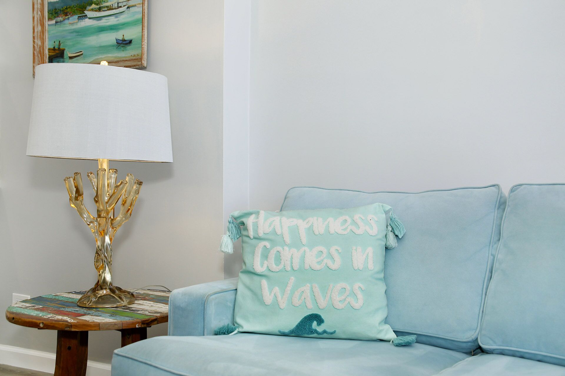 Una habitación con un sofá azul claro, una mesita redonda con una lámpara y un cojín con el texto 'Happiness Comes in Waves'.