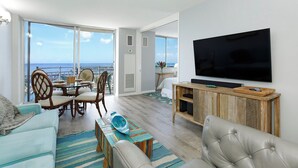 Ein modernes Wohnzimmer mit großem Fernseher, bequemen Sitzgelegenheiten, einem Essbereich und einem Balkon mit Meerblick.